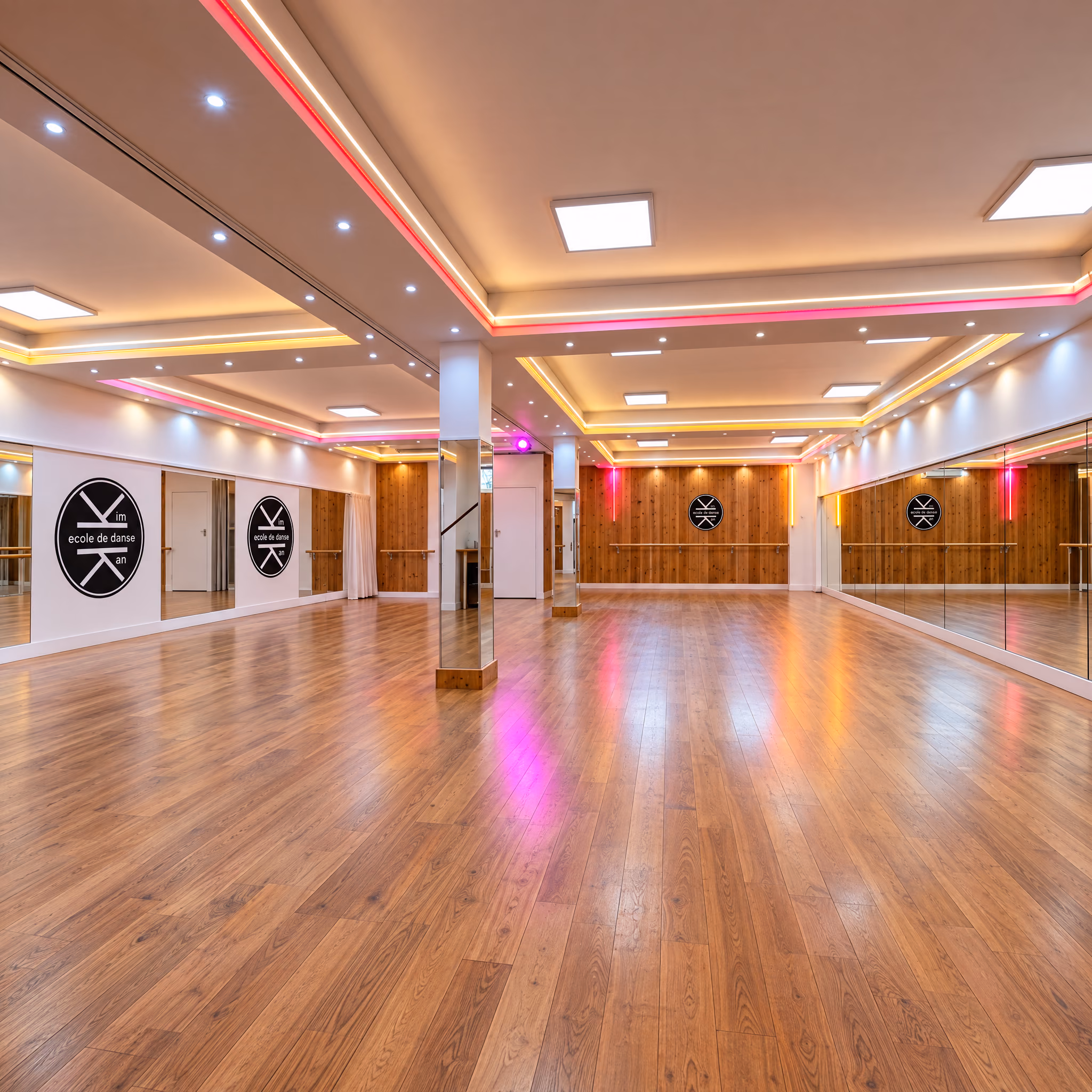 Grande salle de 140 m² de l’Espace Danse Gambetta, vide : murs blancs aux deux médaillons du logo, pilier miroir, lambris et miroirs au fond, parquet sous une lumière rose.