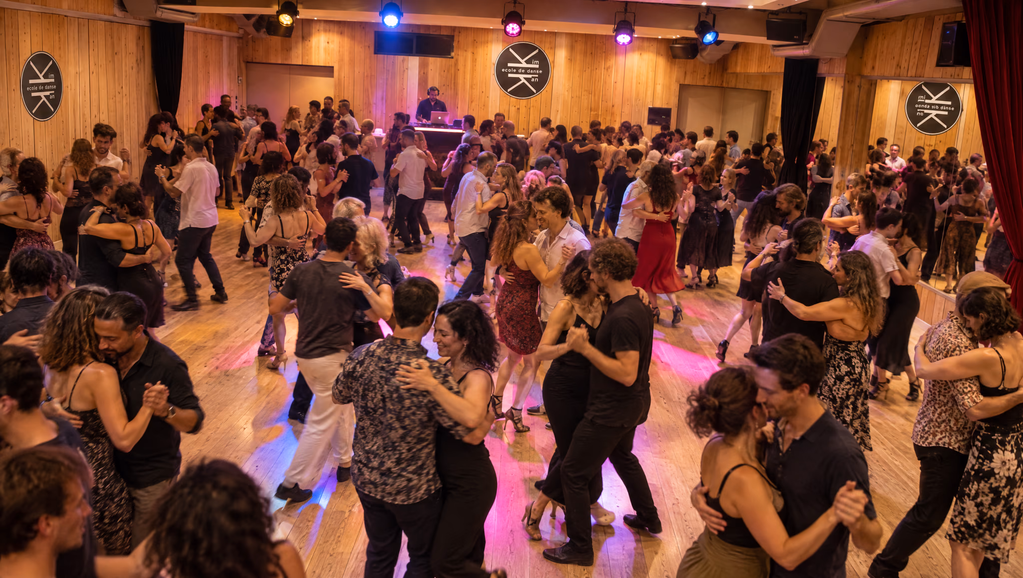 Soirée dansante au Studio des Rigoles vue en plongée : couples sur le parquet sous les projecteurs colorés, DJ au fond, lambris, rideau rouge et miroirs.
